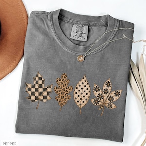 May include: A charcoal gray short-sleeve t-shirt featuring a graphic of four autumn leaves with distinct patterns: a black and tan checkerboard, a brown leopard print, black polka dots, and black flowers. The shirt has a Comfort Colors label.