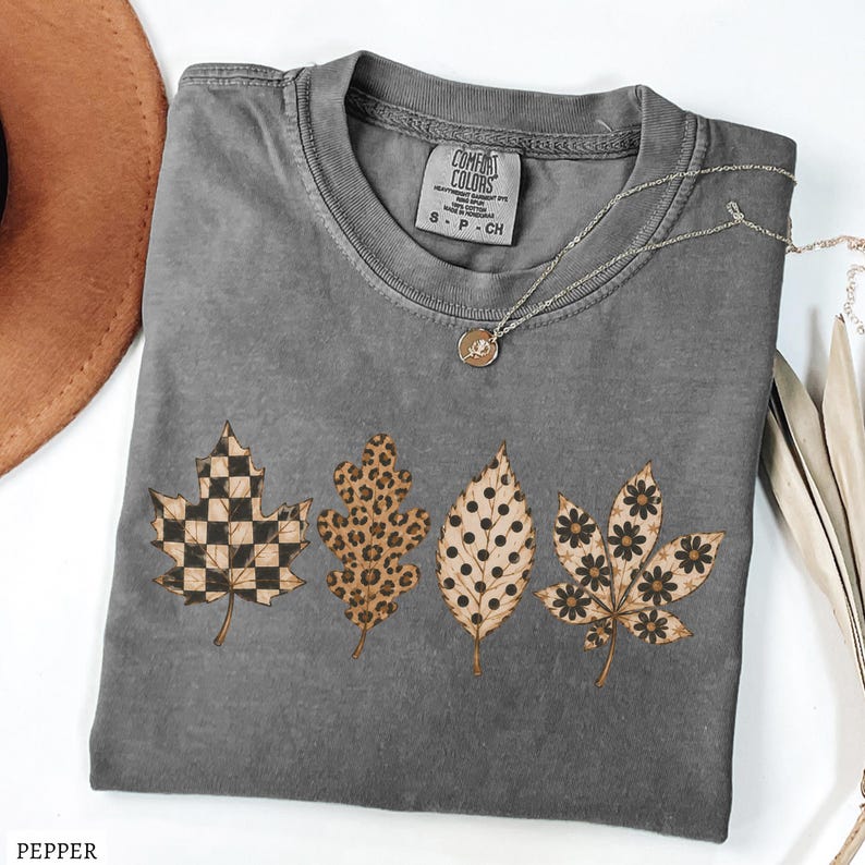 May include: A charcoal gray short-sleeve t-shirt featuring a graphic of four autumn leaves with distinct patterns: a black and tan checkerboard, a brown leopard print, black polka dots, and black flowers. The shirt has a Comfort Colors label.