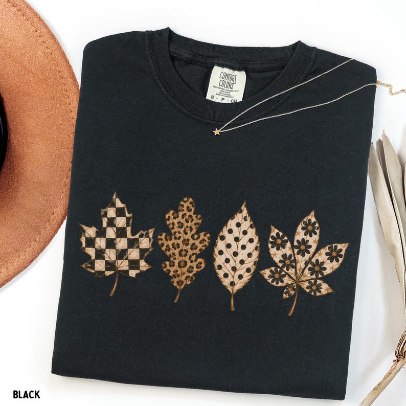 May include: A black crewneck t-shirt featuring a graphic of four autumn leaves arranged in a row. The leaves display distinct patterns including a black and white checkerboard, a brown leopard print, black polka dots, and black floral motifs.