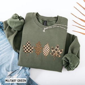 May include: A military green crewneck sweatshirt featuring a graphic print of four autumn leaves. The leaves display distinct patterns including a black and tan checkerboard, a brown leopard print, black polka dots, and black floral motifs.