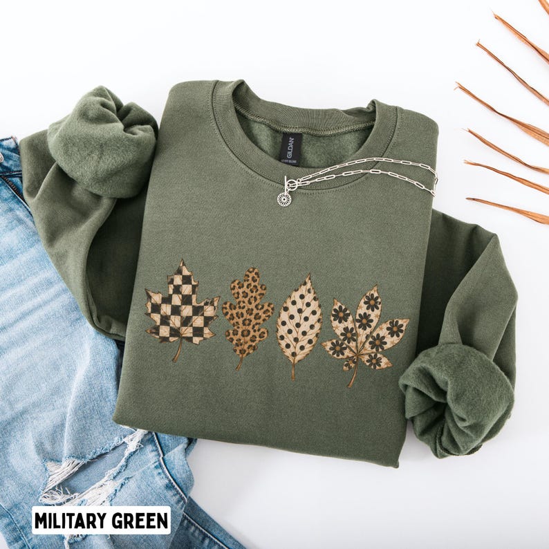 May include: A military green crewneck sweatshirt featuring a graphic print of four autumn leaves. The leaves display distinct patterns including a black and tan checkerboard, a brown leopard print, black polka dots, and black floral motifs.