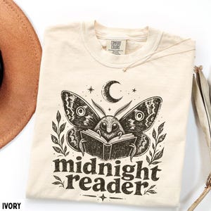 May include: An ivory t-shirt featuring a black graphic print of a moth reading an open book beneath a crescent moon. The design includes leafy branches on both sides and the text "midnight reader" in a bold, dark font below the illustration.