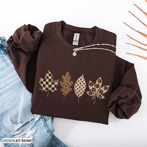 May include: A chocolate brown crewneck sweatshirt featuring a graphic print of four distinct leaves arranged horizontally. The leaves display a checkered pattern, a leopard print, a polka dot design, and a floral pattern in shades of brown and tan.