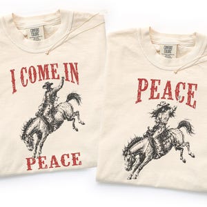 May include: Two ivory t-shirts featuring a distressed red and black graphic print. The left shirt displays a cowboy riding a bucking horse with the text I COME IN PEACE. The right shirt shows a cowgirl on a bucking horse with the text PEACE.