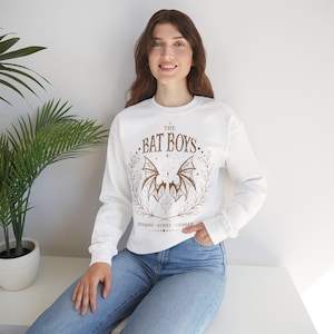 May include: White long-sleeve sweatshirt with a brown graphic of bat wings, the text "THE BAT BOYS," and the names Rhysand, Azriel, and Cassian. The design is surrounded by a wreath and celestial elements.