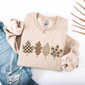 May include: A sand-colored crewneck sweatshirt featuring a graphic print of four distinct leaves arranged in a row. The leaves display various patterns including a black and tan checkerboard, a brown leopard print, black polka dots, and floral.