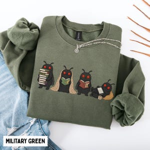 Puede incluir: Una sudadera de cuello redondo color verde militar con un gr&aacute;fico de cuatro criaturas negras con ojos rojos similares a polillas. Una sostiene una pila de libros, otra lee envuelta en una manta, otra usa gafas y otra est&aacute; acostada con una tableta.