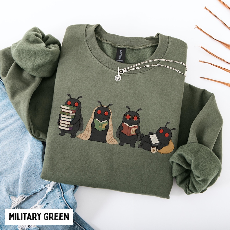 Puede incluir: Una sudadera de cuello redondo color verde militar con un gr&aacute;fico de cuatro criaturas negras con ojos rojos similares a polillas. Una sostiene una pila de libros, otra lee envuelta en una manta, otra usa gafas y otra est&aacute; acostada con una tableta.