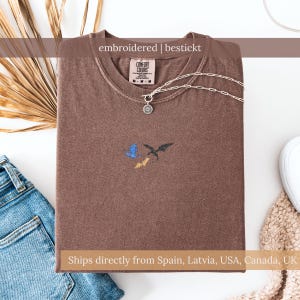 Könnte beinhalten: Braunes T-Shirt mit gesticktem Drachendesign. Das Shirt hat einen Rundhalsausschnitt und ein Comfort Colors-Etikett. Eine silberne Halskette mit einem runden Anhänger befindet sich auf dem Shirt. Der Text "embroidered | bestickt" steht oben.