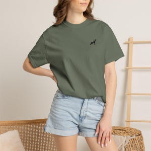 May include: An olive green t-shirt with a small black embroidered design on the upper left chest. The shirt has short sleeves and a crew neck. The model is wearing light blue denim shorts.