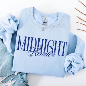 Könnte beinhalten: Hellblaues Sweatshirt mit dem navyblauen Schriftzug "MIDNIGHT Reader". Eine silberne Halskette mit einem Sonnenblumenanhänger liegt auf dem Shirt. Das Sweatshirt ist auf einer Jeans gefaltet.
