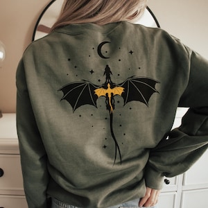 Op de afbeelding: Een groen sweatshirt met een zwart draken silhouet en een gele draak binnen de vleugels. De draak vliegt tegen een achtergrond van een halve maan en sterren.