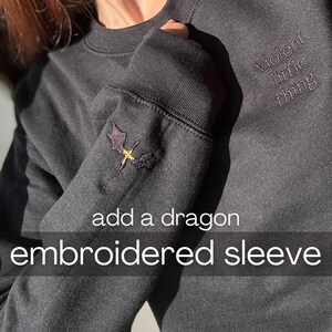May include: Dark gray sweatshirt with embroidered dragon on the sleeve. The text "violent little thing" is embroidered on the upper right chest. The sleeve also has the text "add a dragon embroidered sleeve".