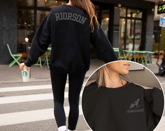 May include: Black crew neck sweatshirt with the word "RIORSON" arched across the upper back. A smaller image shows the front of the sweatshirt with a dragon graphic and the word "WINGLEADER". The sweatshirt is worn with black leggings and a black cap.