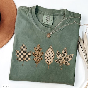 May include: A moss green t-shirt featuring a graphic print of four distinct leaves. Each leaf displays a different pattern: a black and tan checkerboard, a brown leopard print, black polka dots, and a black floral design on a tan background.