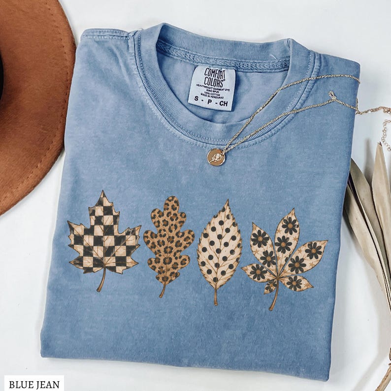 May include: A blue jean colored t-shirt with a Comfort Colors label at the neck. The shirt features a graphic print of four leaves, each with a different pattern: a black and tan checkerboard, a brown leopard print, black polka dots, and black flowers.