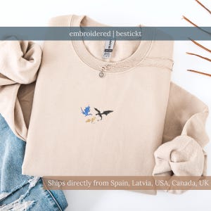 Könnte beinhalten: Beiges Sweatshirt mit Rundhalsausschnitt und gestickten Drachendesigns in Blau, Schwarz und Gelb. Der Text "embroidered | bestickt" befindet sich über dem Ausschnitt. Eine silberne Halskette mit Sonnenanhänger befindet sich auf dem Sweatshirt.