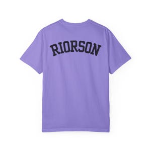 May include: A lilac t-shirt featuring the word "RIORSON" in black, arched lettering on the back. The short-sleeved shirt has a crew neck. The fabric is a soft material.