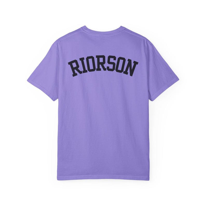 May include: A lilac t-shirt featuring the word "RIORSON" in black, arched lettering on the back. The short-sleeved shirt has a crew neck. The fabric is a soft material.