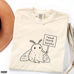 May include: An ivory-colored t-shirt featuring a black line-art illustration of a fluffy moth holding a sign that reads "read more books." The shirt has a crew neckline and a Comfort Colors brand label printed on the inside of the collar.