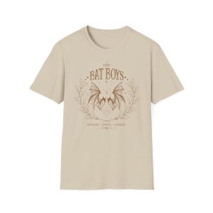 May include: Beige t-shirt featuring a brown graphic of a bat with spread wings, surrounded by a laurel wreath. The text "THE BAT BOYS" is above the bat, with names below. A casual, short-sleeved tee.