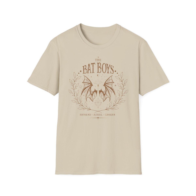 May include: Beige t-shirt featuring a brown graphic of a bat with spread wings, surrounded by a laurel wreath. The text "THE BAT BOYS" is above the bat, with names below. A casual, short-sleeved tee.