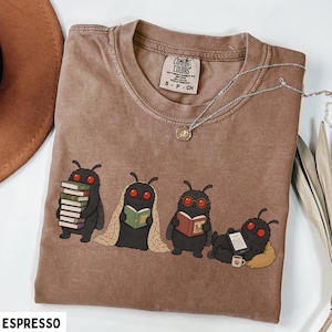 Puede incluir: Una camiseta marr&oacute;n con un gr&aacute;fico de cuatro criaturas negras parecidas a polillas con ojos rojos leyendo libros y usando un lector electr&oacute;nico. La camiseta tiene una etiqueta Comfort Colors en el cuello y un collar de cadena plateada.