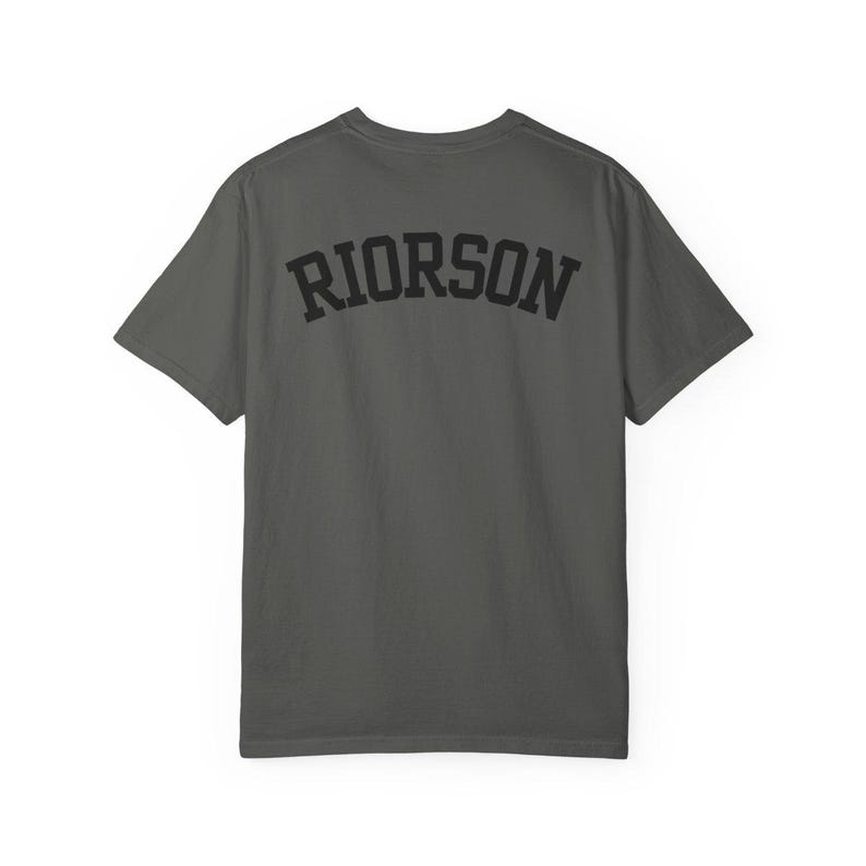 May include: A dark grey t-shirt featuring the word "RIORSON" in black, arched lettering across the back. The shirt has a crew neck and short sleeves.