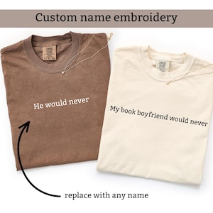Può includere: Due magliette, una marrone e una color crema, con testo. La maglietta marrone dice "He would never", e la maglietta color crema dice "My book boyfriend would never". Entrambe le magliette hanno una collana d'oro. Il testo "Custom name embroidery" è in alto.