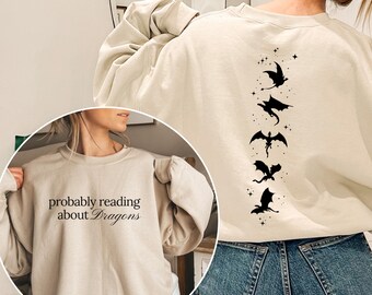 Puede incluir: Sudadera beige con el texto "probably reading about Dragons" en la parte delantera. La parte trasera presenta un diseño vertical de siluetas de dragones negros y estrellas. La sudadera es de estilo casual y cómodo.