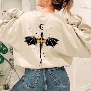 Könnte beinhalten: Beigefarbenes Sweatshirt mit einem schwarzen Drachendesign mit gelber Brust, schwarzen Flügeln und einem Halbmond und Sternen. Der Drache ist auf der Rückseite zentriert. Das Sweatshirt hat lange Ärmel und einen Rundhalsausschnitt.