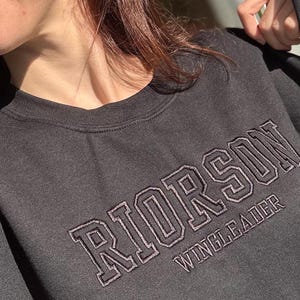 Könnte beinhalten: Ein dunkelgraues Sweatshirt mit dem Wort "RIORSON" in einem College-Schriftzug auf der Brust, darunter "WINGLEADER". Das Sweatshirt hat einen Rundhalsausschnitt und lange Ärmel.