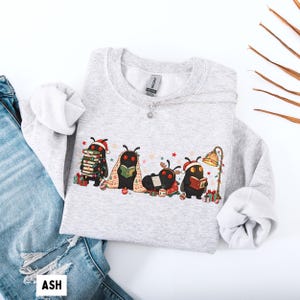 May include: A light gray crewneck sweatshirt featuring a graphic of four black moth-like creatures with red eyes. They are wearing Santa hats, reading books, and sitting among Christmas gifts, a patterned blanket, and a floor lamp.