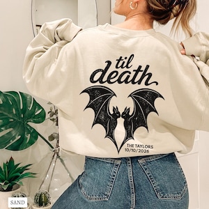 May include: A sand-colored crewneck sweatshirt featuring a black graphic on the back. The design displays two bats facing each other with wings spread, the text "til death" above them, and "THE TAYLORS 10/10/2026" printed in black below the bats.