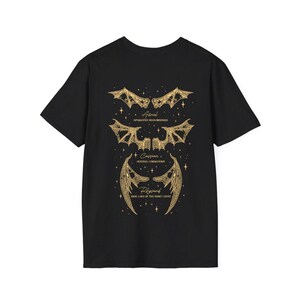 May include: Black t-shirt featuring gold-colored illustrations of bat-like wings and text. The text includes names and titles: Azriel, Spymaster Shadowsinger; Cassian, General Commander; and Rhysand, High Lord of the Night Court.