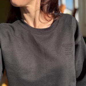 Może przedstawiać: Ciemnoszara bluza z haftowanym napisem "violent little thing" na piersi. Bluza z okrągłym dekoltem jest wykonana z miękkiego materiału i ma długie rękawy.