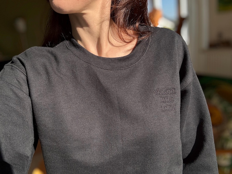 Może przedstawiać: Ciemnoszara bluza z haftowanym napisem "violent little thing" na piersi. Bluza z okrągłym dekoltem jest wykonana z miękkiego materiału i ma długie rękawy.