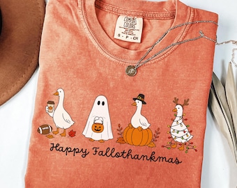 Può includere: Una t-shirt a maniche corte color corallo con una grafica di quattro oche in vari costumi, tra cui un fantasma, un pellegrino e una avvolta da luci natalizie. Il testo "Happy Fallothankmas" è stampato sotto. Etichetta Comfort Colors presente.