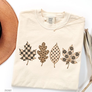 May include: An ivory-colored crew neck t-shirt featuring a graphic print of four distinct autumn leaves arranged in a row. The leaves display varied patterns including a black and ivory checkerboard, a brown leopard print, black polka dots, and black flowers.