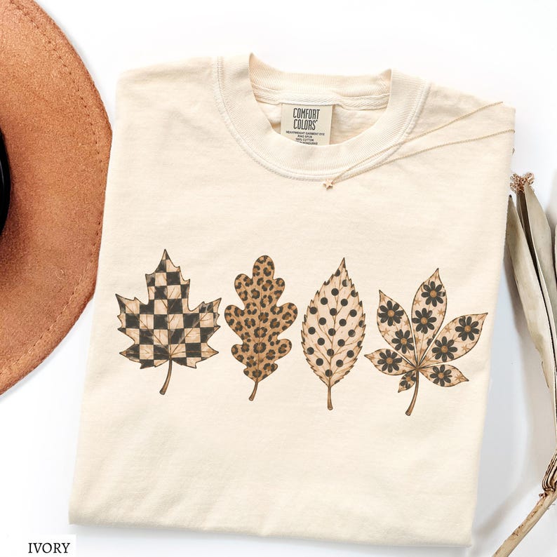 May include: An ivory-colored crew neck t-shirt featuring a graphic print of four distinct autumn leaves arranged in a row. The leaves display varied patterns including a black and ivory checkerboard, a brown leopard print, black polka dots, and black flowers.