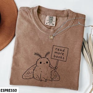 May include: An espresso-colored short-sleeve t-shirt featuring a black line-art illustration of a fuzzy moth holding a sign that reads read more books. The shirt has a crew neckline and a Comfort Colors brand label inside the collar.