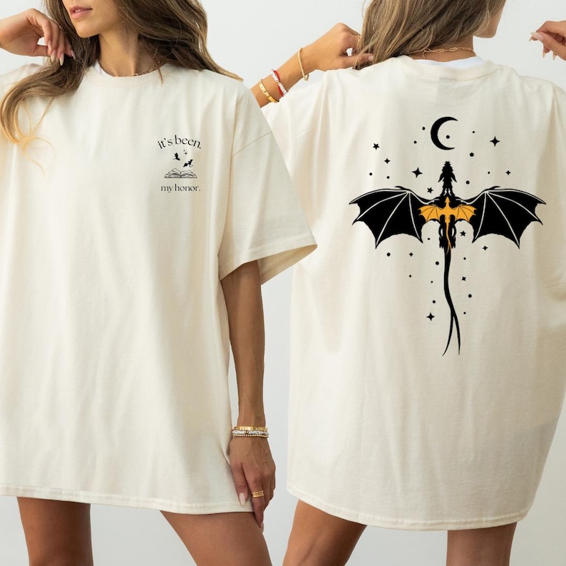 May include: Cream-coloured t-shirt with front and back graphics. The front has text and a book graphic. The back shows a dragon with black wings, a crescent moon, and stars. A casual, oversized fit.