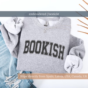 Könnte beinhalten: Ein graues Sweatshirt mit dem Wort "BOOKISH" in schwarzen Blockbuchstaben gestickt. Das Sweatshirt hat einen Rundhalsausschnitt und lange Ärmel. Eine silberne Halskette mit einem kleinen Anhänger ist darüber drapiert. Der Text "embroidered | bestickt" ist darüber.