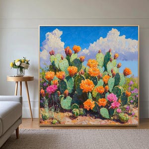 以下が含まれることがあります： 砂漠の風景を描いた正方形の額入り絵画。緑色のウチワサボテンにはオレンジとピンクの花が咲き、白い雲が浮かぶ青い空を背景にしています。鮮やかでカラフルなアート作品です。