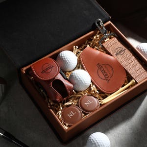 May include: A golf gift set in a brown box, including golf balls, a leather scorecard holder, a golf glove holder, a keychain, and ball markers. The items have the name "NEWELL" printed on them. The box also contains golf tees.
