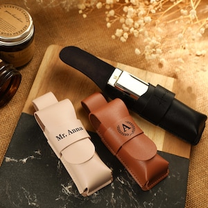 Puede incluir: Una variedad de estuches de cuero y una maquinilla de afeitar negra. Dos estuches son de color rosa claro y marrón, con el texto "Mr. Anna" y una inicial "A" en una corona. También hay una maquinilla de afeitar de cuero negro. Los artículos están sobre una tabla de cortar de madera.