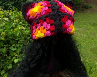 May include: A colorful crochet hat with cat ears. The hat features a black border with squares of red, pink, yellow, and orange. The hat is displayed on a black wig. The background is green foliage.