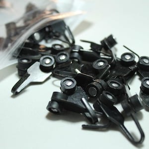 May include: A pile of small, black metal curtain hooks with rollers and silver rivets. The hooks are spread on a white surface, some still in a clear plastic bag. These hooks are designed for hanging curtains.