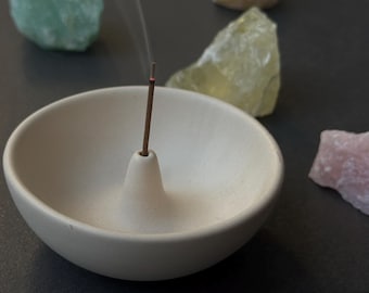 May include: A white ceramic incense holder with a small, round opening in the center. A single incense stick is burning in the holder, with smoke rising from the top. There are four small, colorful stones surrounding the holder.