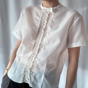 Könnte beinhalten: Eine weiße Bluse mit kurzen Ärmeln und einem Spitzenpanel auf der Vorderseite.